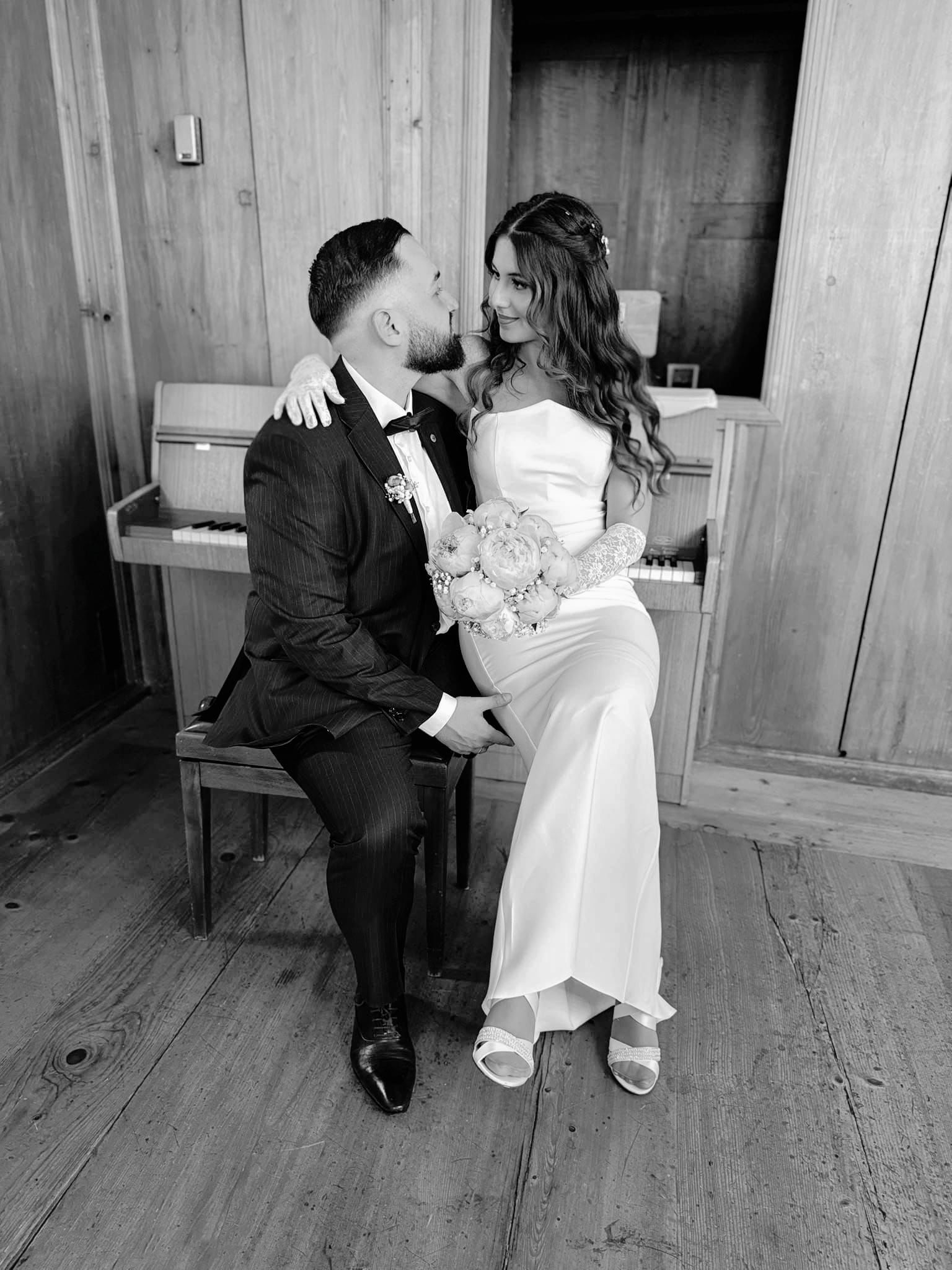 Schwarz-weiss Foto von Ilayda und Tarkan im Hochzeitsoutfit, sitzend vor einem alten Klavier mit Brautstrauss